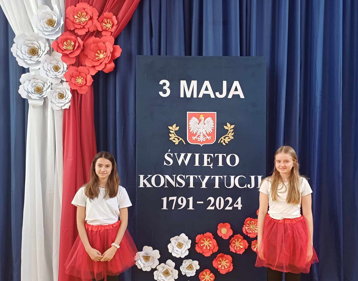 dwie dziewczynki w&nbsp;galowych strojach na&nbsp;tle planszy z&nbsp;napisem Konstytucja 3 Maja