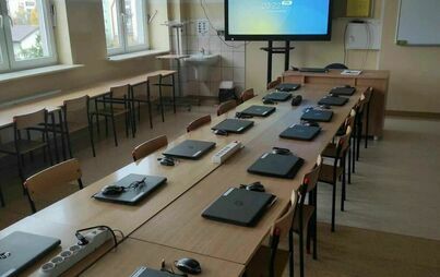 pracownia komputerowa z rozłożonymi laptopami, przy biurku nauczyciela stoi interaktywny monitor.