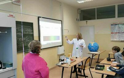 sala lekcyjna, uczniowie siedzą w ławkach. Nauczycielka chemii pokazuje w kierunku tablicy interaktywnej. Na pierwszej ławce model kuli ziemskiej i przyrządy labolatoryjne.